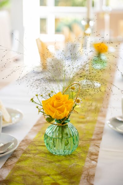 Ein Glas mit einer gelben Blume steht als Dekoration auf einem langen Tisch